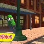Animated green cucumber character peeks from behind a lamppost on a sunny brick sidewalk, VeggieTales logo in the corner.