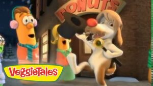 Animated VeggieTales scene: a white dog with a red nose rings a bell while a carrot character watches nearby near a building with VeggieTales signage in the background.