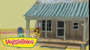 VeggieTales animation: a small brown house with a blue-green metal roof on a yellow street, two veggie characters stand in front (green cucumber with a blue hat and yellow character with sunglasses).