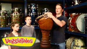 Man shows a large wooden drum to two kids in a music store; VeggieTales logo appears in the corner.