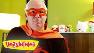 Man wearing a red superhero mask and orange cape in a room with VeggieTales logo foregrounded on the bottom left.