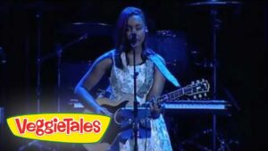 Female singer performing on stage, playing an acoustic guitar and singing into a mic, VeggieTales logo visible in the corner.