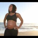 Pregnant woman in a black sports bra and leggings standing on a beach with the ocean in the background, smiling. (informative)