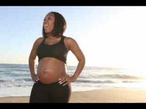 Pregnant woman in a black sports bra and leggings standing on a beach with the ocean in the background, smiling. (informative)