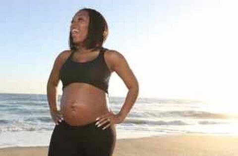 Pregnant woman in a black sports bra and leggings standing on a beach with the ocean in the background, smiling. (informative)