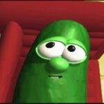 Green pickle character with large eyes and a small smile, lounging on a red couch in a bright room.