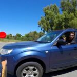 Man in a blue SUV aims a red dart at a large outdoor dartboard in a parking lot with a clear blue sky.