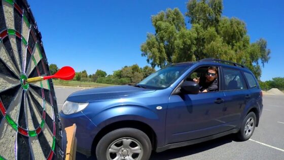 Man in a blue SUV aims a red dart at a large outdoor dartboard in a parking lot with a clear blue sky.