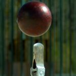 Red cricket ball suspended in mid-air above a metal stump, with a green fence background, conveying a wicket action moment.