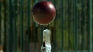 Red cricket ball suspended in mid-air above a metal stump, with a green fence background, conveying a wicket action moment.