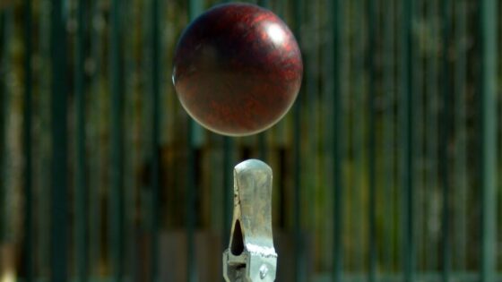 Red cricket ball suspended in mid-air above a metal stump, with a green fence background, conveying a wicket action moment.