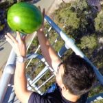 Person on a metal railing high above ground reaching with both hands to place a large green watermelon on the railing.