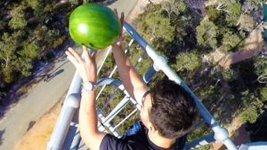 Person on a metal railing high above ground reaching with both hands to place a large green watermelon on the railing.