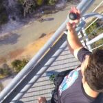 Person standing on a tall metal railing on a tower, holding a rock toward the edge with a ground path visible below.