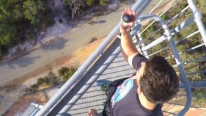 Person standing on a tall metal railing on a tower, holding a rock toward the edge with a ground path visible below.