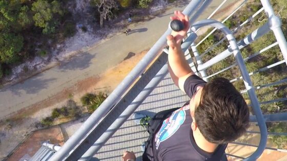 Person standing on a tall metal railing on a tower, holding a rock toward the edge with a ground path visible below.