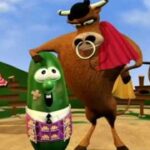 Cartoon bull wearing a red cape stands beside a cheerful green vegetable character in a field scene with spectators.