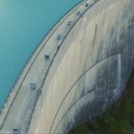 A large curved concrete dam holding back a turquoise lake, with a grassy hillside on the right and a road along the dam's crest visible.