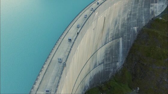 A large curved concrete dam holding back a turquoise lake, with a grassy hillside on the right and a road along the dam's crest visible.