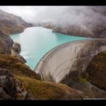 Curved concrete dam with a turquoise reservoir and misty mountains in the background.