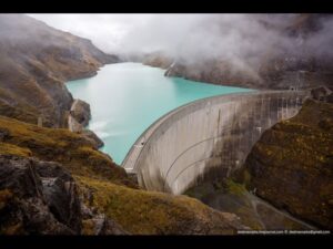 Curved concrete dam with a turquoise reservoir and misty mountains in the background.