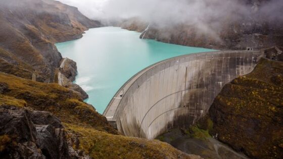 Curved concrete dam with a turquoise reservoir and misty mountains in the background.