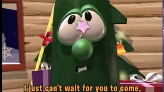 Cartoon green creature dressed as a Christmas tree with presents around, looking toward the viewer.