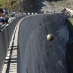Young man stands on a bridge railing inspecting a black-and-yellow ball rolling down a tall concrete dam wall near a rocky mountainside.