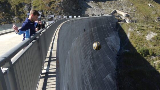 Young man stands on a bridge railing inspecting a black-and-yellow ball rolling down a tall concrete dam wall near a rocky mountainside.