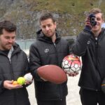 Three men in matching black jackets pose outdoors on a seaside boardwalk, each holding different sports balls (football, soccer ball, basketball, tennis balls).