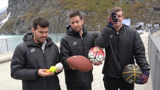 Three men in matching black jackets pose outdoors on a seaside boardwalk, each holding different sports balls (football, soccer ball, basketball, tennis balls).