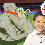 Child scientist wearing a bow tie and yellow glasses smiles beside a banana-skeleton dinosaur on a green field set with a colorful backdrop and 'Imagine That' sign nearby.