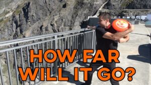 Man at a cliff overlook throws a frisbee while holding a selfie stick; large orange text reads 'HOW FAR WILL IT GO?'