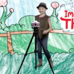 Girl posing with a camera on a tripod in front of a colorful drawn backdrop that says 'Imagine That!' in a playful scene.