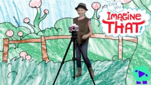 Girl posing with a camera on a tripod in front of a colorful drawn backdrop that says 'Imagine That!' in a playful scene.