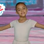 Young girl in a white leotard with arms extended, posing in front of a colorful mural reading 'Imagine That!' with pink hearts nearby.