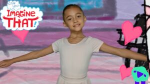 Young girl in a white leotard with arms extended, posing in front of a colorful mural reading 'Imagine That!' with pink hearts nearby.