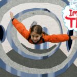 Child in an orange jacket flying through spiraling blue-gray tunnel walls in an indoor set, with 'Imagine That!' logo at the top-right.