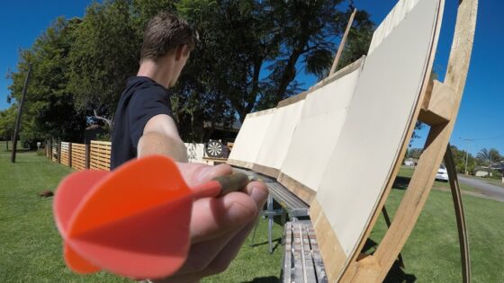Person extending their arm toward the camera, holding an orange plastic dart while standing beside a curved wooden frame with white panels outdoors on a sunny day.