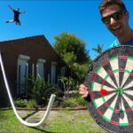Man in a blue shirt holds a giant dartboard outdoors while another person jumps mid-air in the background in a sunny yard.