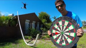 Man in a blue shirt holds a giant dartboard outdoors while another person jumps mid-air in the background in a sunny yard.