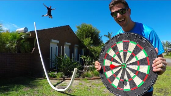 Man in a blue shirt holds a giant dartboard outdoors while another person jumps mid-air in the background in a sunny yard.