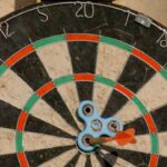 Dartboard with a blue three-armed spinner at the center and an orange dart stuck near the bullseye.