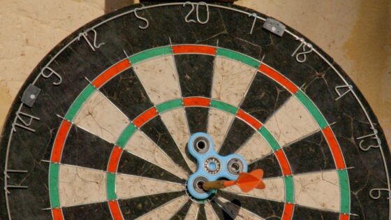 Dartboard with a blue three-armed spinner at the center and an orange dart stuck near the bullseye.