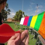 Person holding a red plastic target paddle close to the camera as colorful boards are set up on stands in a park-like field.