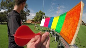 Person holding a red plastic target paddle close to the camera as colorful boards are set up on stands in a park-like field.