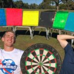 Two men pose outdoors with a row of colorful boards and a large dartboard between them, smiling.",