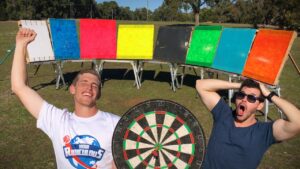Two men pose outdoors with a row of colorful boards and a large dartboard between them, smiling.",