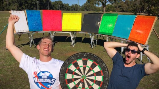 Two men pose outdoors with a row of colorful boards and a large dartboard between them, smiling.",