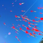 Blue sky with many red-tipped darts flying toward a circular, multicolored target at right; a tall metal tower on the left acts as a backdrop (fairground game).